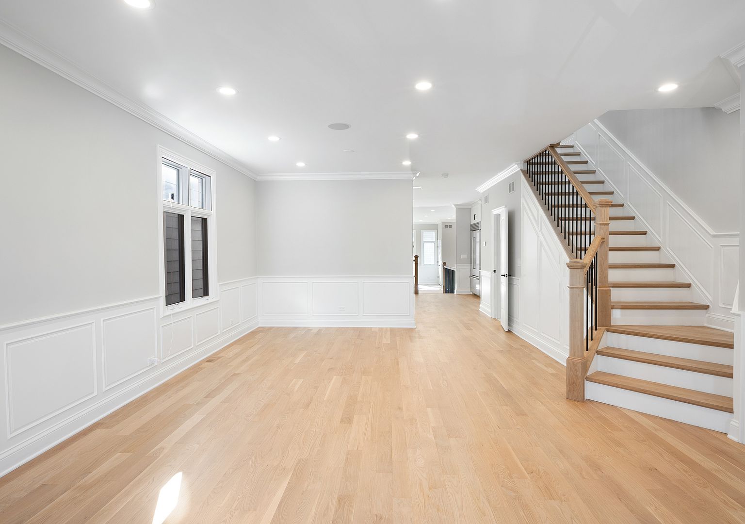 Open great room on the main floor, looking from the formal dining toward the stair landing. White Oak flooring runs the length of the room, with classical wainscoting along the lower walls and a custom staircase in white oak treads and black iron balusters