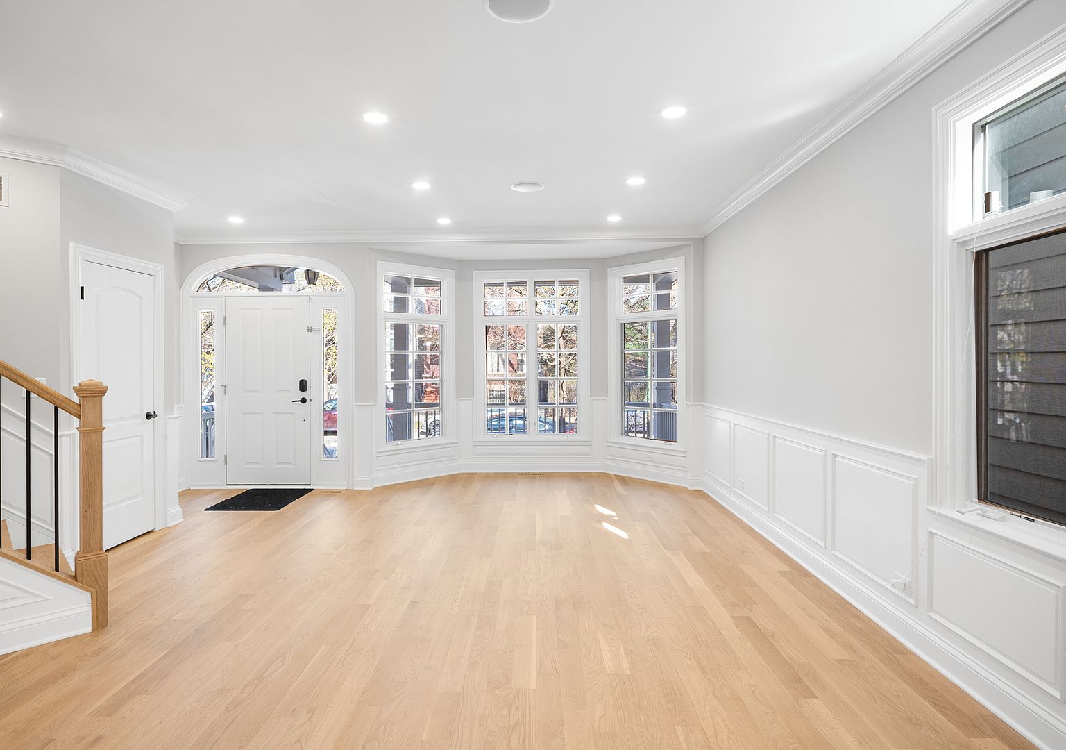 Front foyer with arched-top entry door, sidelights, and a bay window curving outward to the porch. Classical wainscoting wraps the room and recessed ceiling lights illuminate the White Oak floor