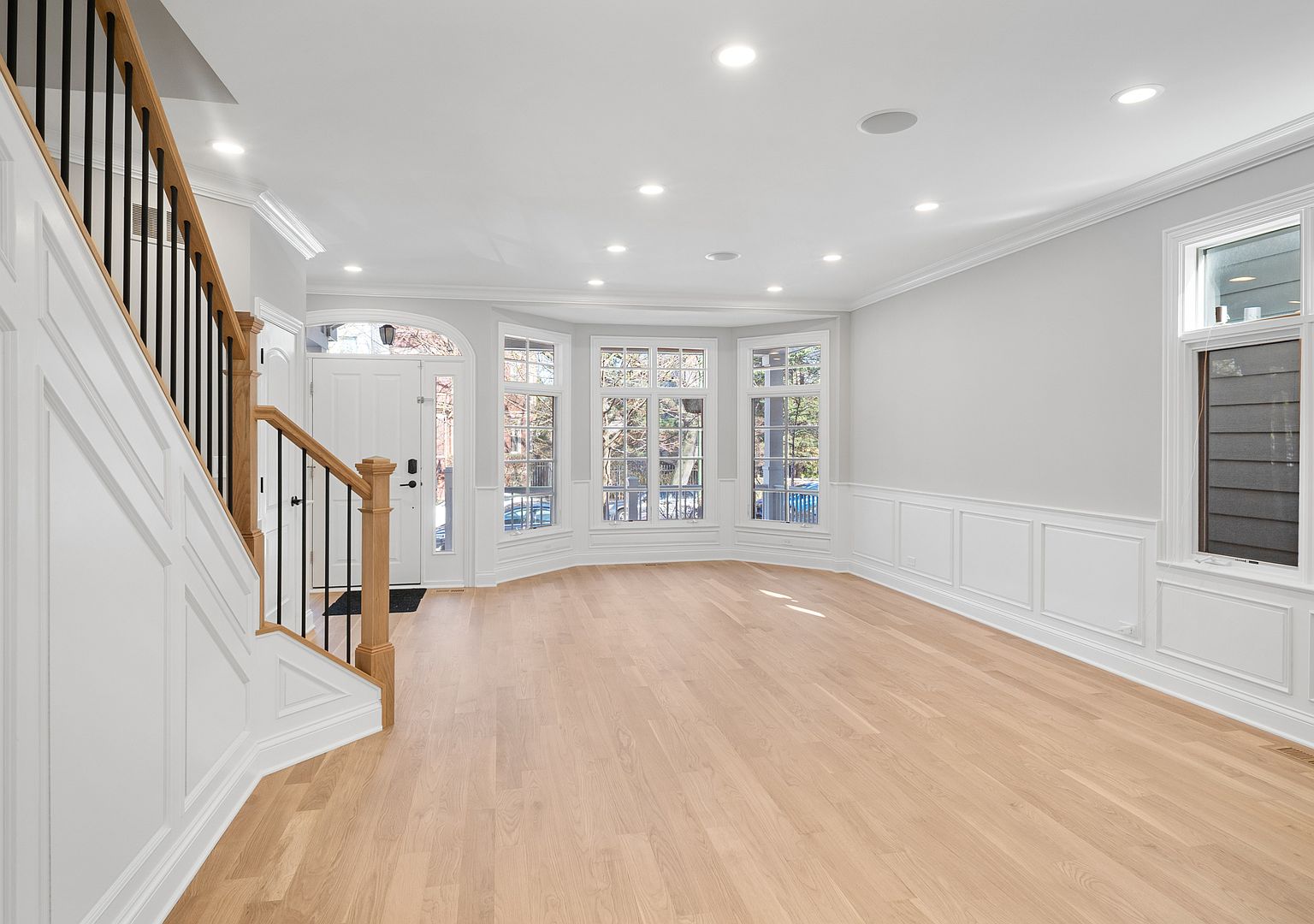 Entry view from the foot of the stairs, looking across the open main floor toward the bay window and front door. Sunlight from the bay falls across the White Oak floor in long stripes